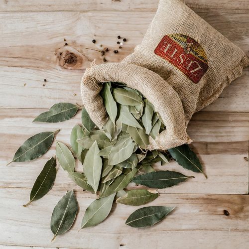 Organic laurel leaves spilling from a burlap sack onto a wooden surface, with peppercorns scattered nearby.